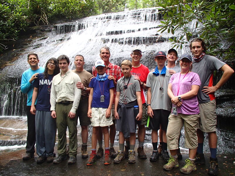 A group of about a dozen people are posing for a photo in front of a waterfall. They appear to be of various ages and are dressed in casual clothing, suitable for hiking or outdoor activities. The waterfall is wide and multi-tiered, creating a scenic backdrop for the group. The overall impression is one of a group outing or excursion in a natural setting.